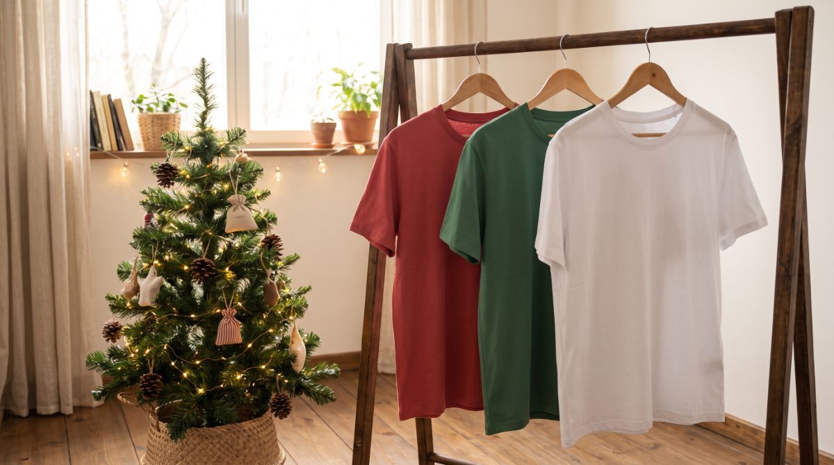 Three plain t-shirts in red, green and white hanging on a wooden rail beside a small decorated Christmas tree