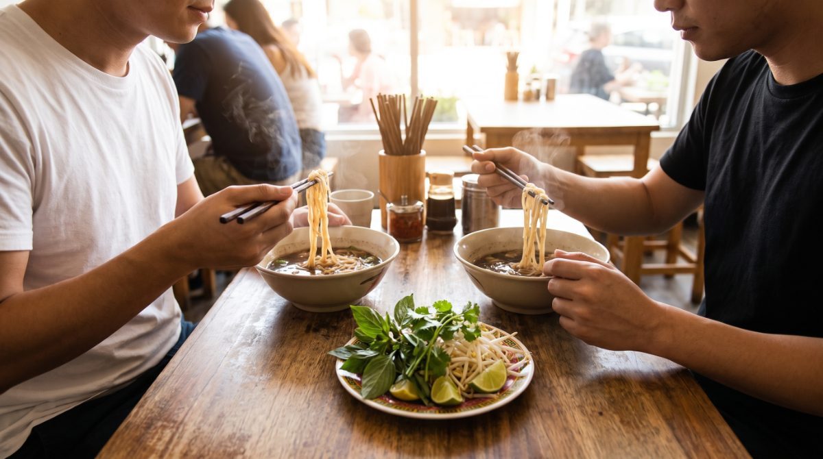 Two friends in plain white and black t-shirts eating bowls of pho with chopsticks at a wooden table in a noodle shop