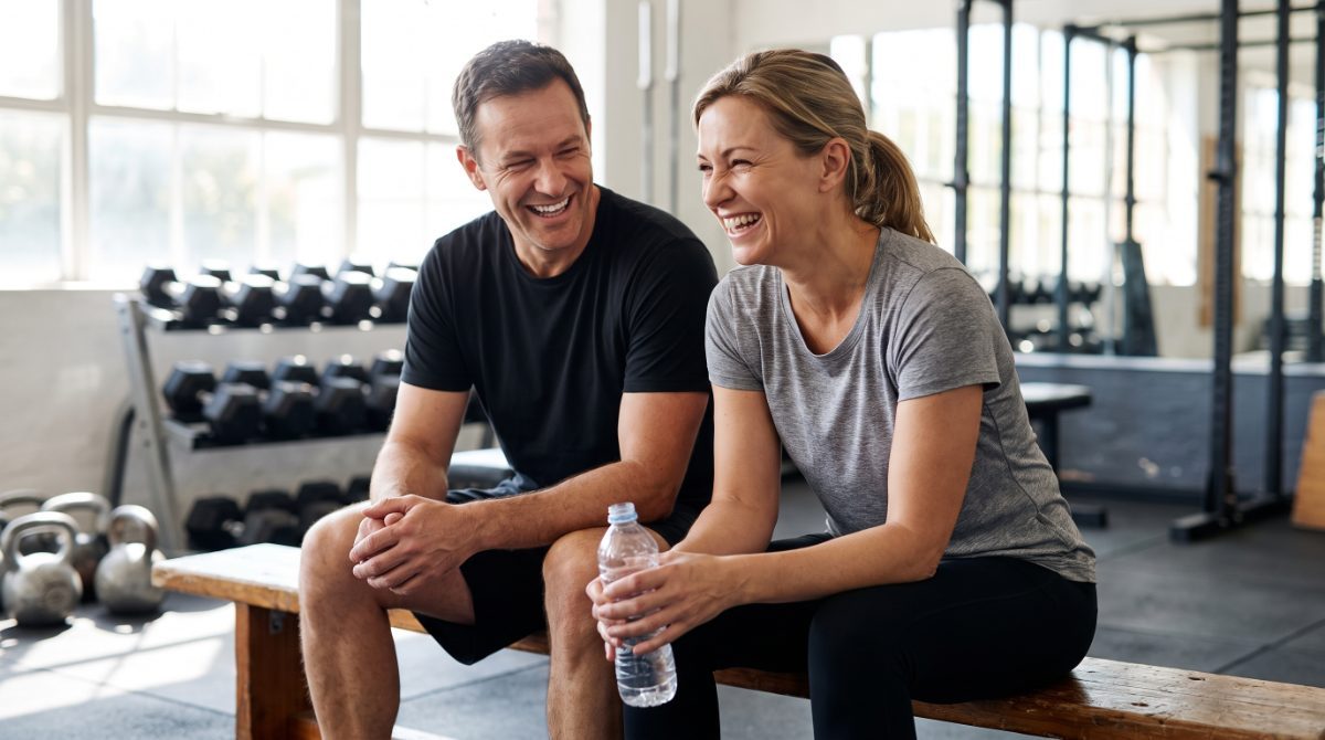 Two friends in plain t-shirts laughing on a gym bench between sets, one holding a water bottle
